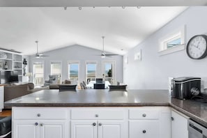 Una cocina moderna con armarios blancos, encimera de granito y una isla integrada. Hay un comedor con mesa y sillas, y un reloj grande en la pared.