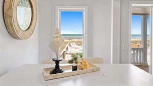 A room with a large window, a wooden wall clock, a table with a tray, and a view of the beach. - Sea2Sky by Carolina Getaway - Oceanfront Home w/ Private Pool & Heated Spa (Kure Beach)