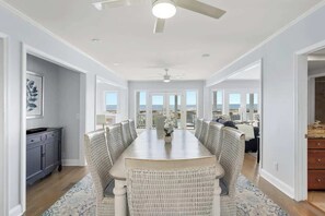 A dining area with a long table and wicker chairs, a ceiling fan, and a view through large windows. - Sea2Sky by Carolina Getaway - Oceanfront Home w/ Private Pool & Heated Spa (Kure Beach)