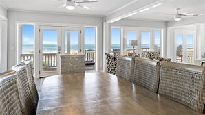 A dining area with a large table and wicker chairs, large windows with ocean views, and ceiling fans. - Sea2Sky by Carolina Getaway - Oceanfront Home w/ Private Pool & Heated Spa (Kure Beach)