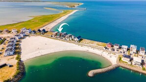 Vue aérienne d’une zone résidentielle côtière, avec des maisons le long de la plage et un phare à l’horizon.
