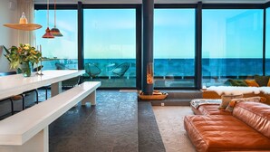 Un salon moderne doté d’une grande fenêtre donnant sur la mer, d’une cheminée et d’un coin repas avec un banc et une table.