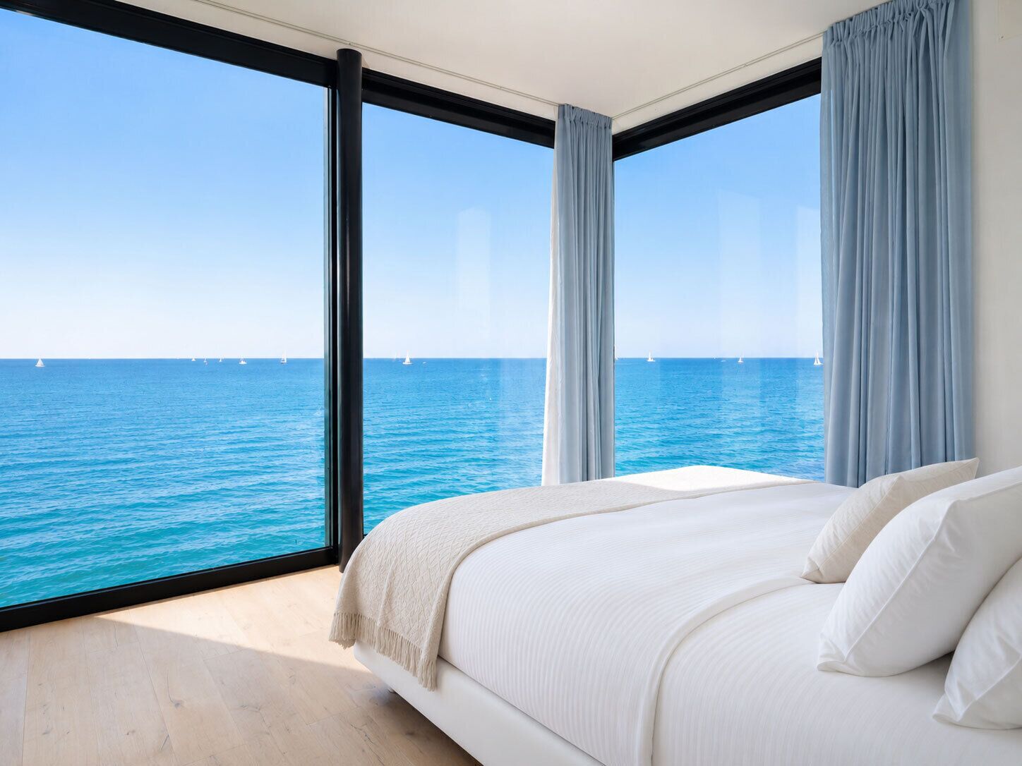 Un dormitorio con un ventanal grande con vistas al mar, una cama con ropa blanca y suelo de madera.