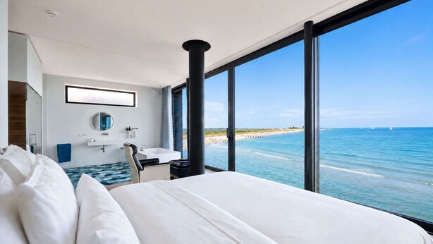 Une chambre d’hôtel moderne avec un grand lit, une salle de bain dotée d’un miroir et d’un lavabo, et une vue sur l’océan.