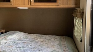 Un intérieur de camping-car compact comprenant un lit, des placards en bois et une fenêtre avec des stores.