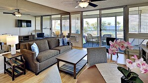 A spacious living room with a sofa, coffee table, and chairs, large windows with a view of the ocean, and a ceiling fan. - Where Turquoise Waters Beckon Kihei Condo Ocean Views Rooftop Garden Pool (Kihei)