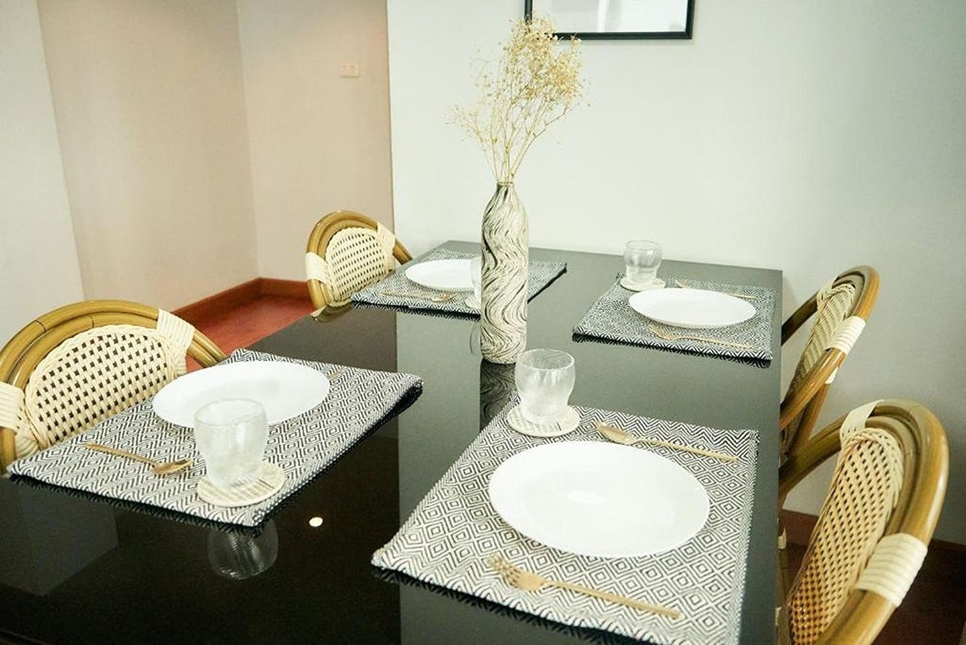 Una mesa de comedor con cuatro sillas, cada una con un mantel individual estampado y un plato blanco. Un jarrón con flores secas está sobre la mesa.