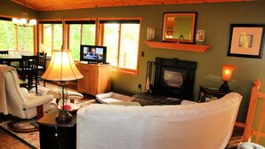 A cozy living room with a fireplace, comfortable seating, a television, and a dining area with natural light from large windows. - Woodland Trails Cabin on 130 acres with wildlife and miles of private trails (Hinckley)