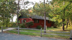 A red cabin with a wooden deck, surrounded by trees and a gravel path. - Woodland Trails Cabin on 130 acres with wildlife and miles of private trails (Hinckley)