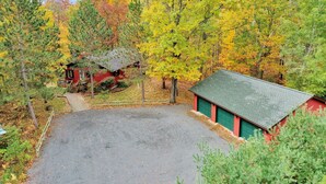 A red cabin with a green garage door, surrounded by a gravel parking area and dense forest with autumn foliage. - Woodland Trails Cabin on 130 acres with wildlife and miles of private trails (Hinckley)