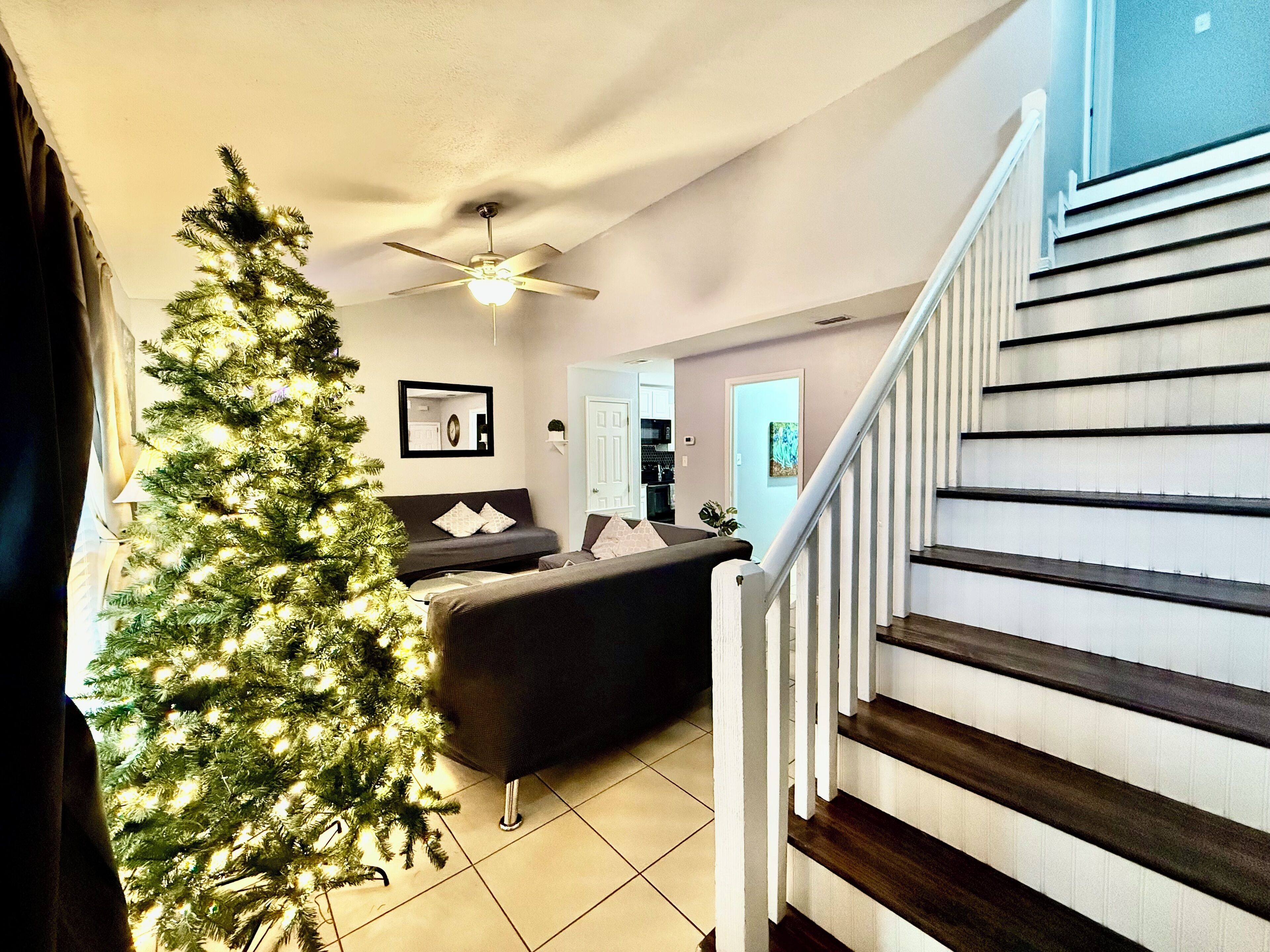 Una sala de estar con un gran árbol de Navidad, un sofá oscuro, un ventilador de techo y una escalera con escalones de madera.