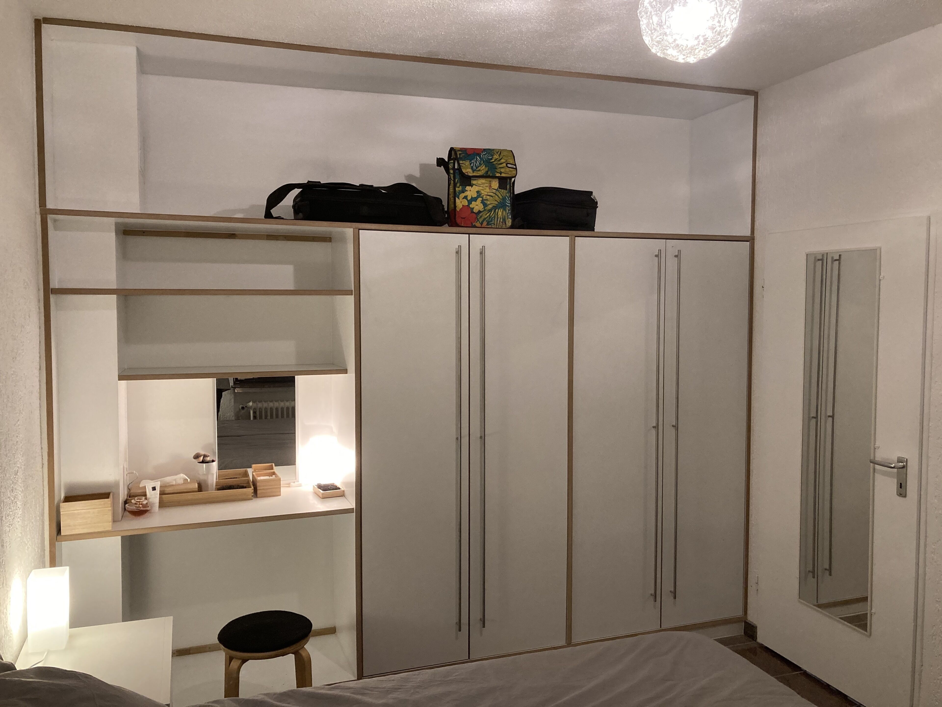 A bedroom with a bed, a desk, a chair, a wardrobe, and a shelf with various items.