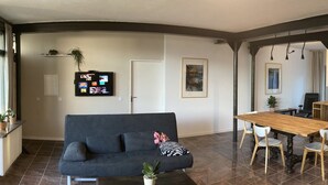 A modern living room with a grey sofa, wooden dining table, and chairs. There is a kitchen area with a stove, sink, and cabinets. - Ferienidylle Haus Königsberg, Child-friendly, Dog-friendly, Barrier-free (Michelstadt)