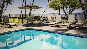 A swimming pool with lounge chairs and umbrellas, surrounded by palm trees and a view of the ocean.