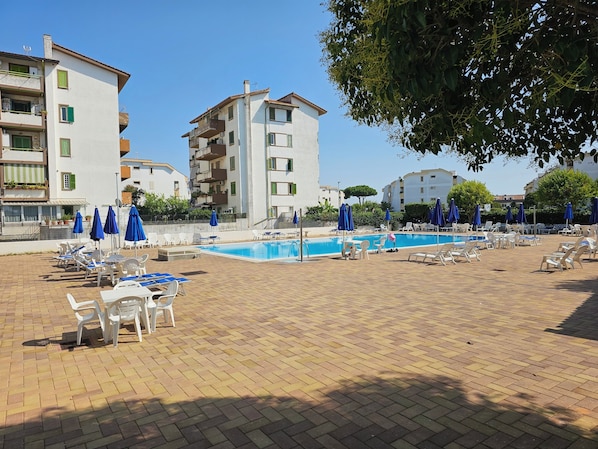 Un área junto a la piscina con sillas de descanso y sombrillas, rodeada de edificios.