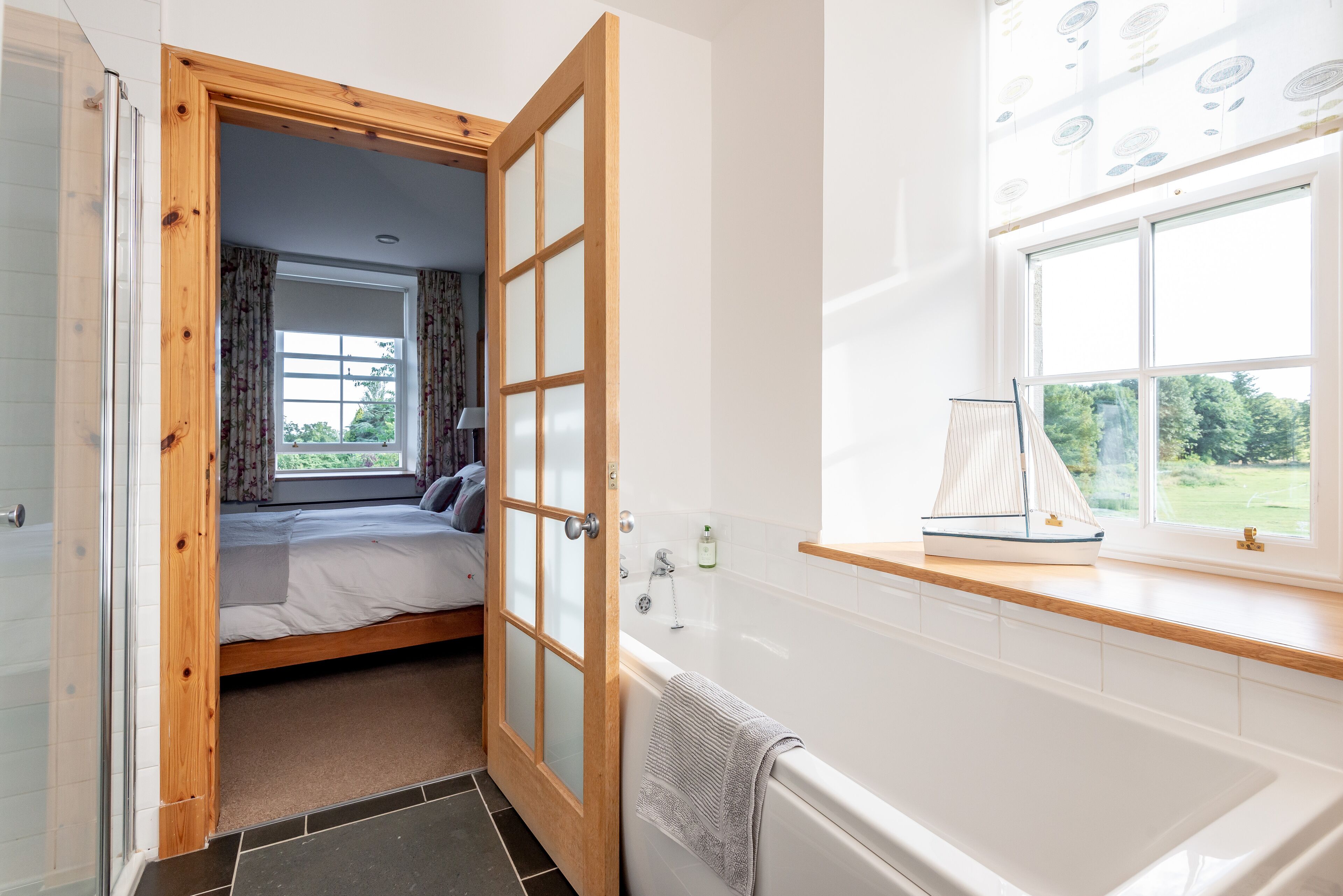 Un dormitorio con puerta de madera, una cama, una ventana con cortinas, una bañera y un peque ño modelo de velero en el alféizar.
