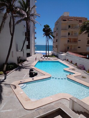 A swimming pool with a view of the ocean, surrounded by palm trees and modern buildings.