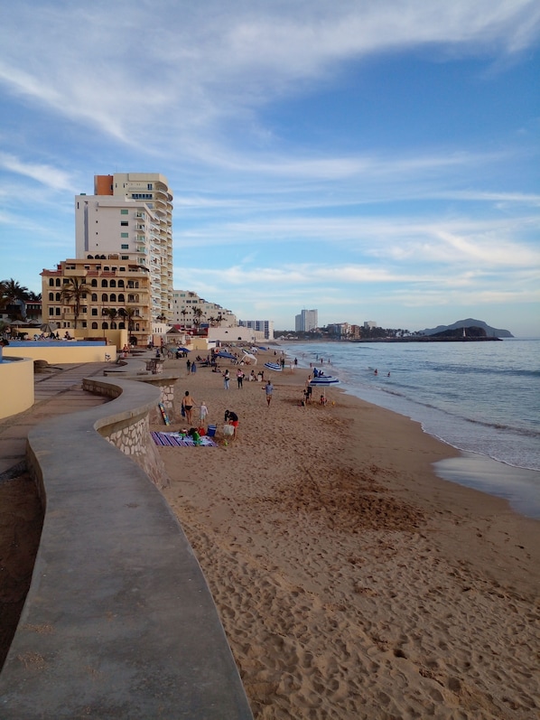 A beach with a curved walkway, people relaxing, and buildings in the background.