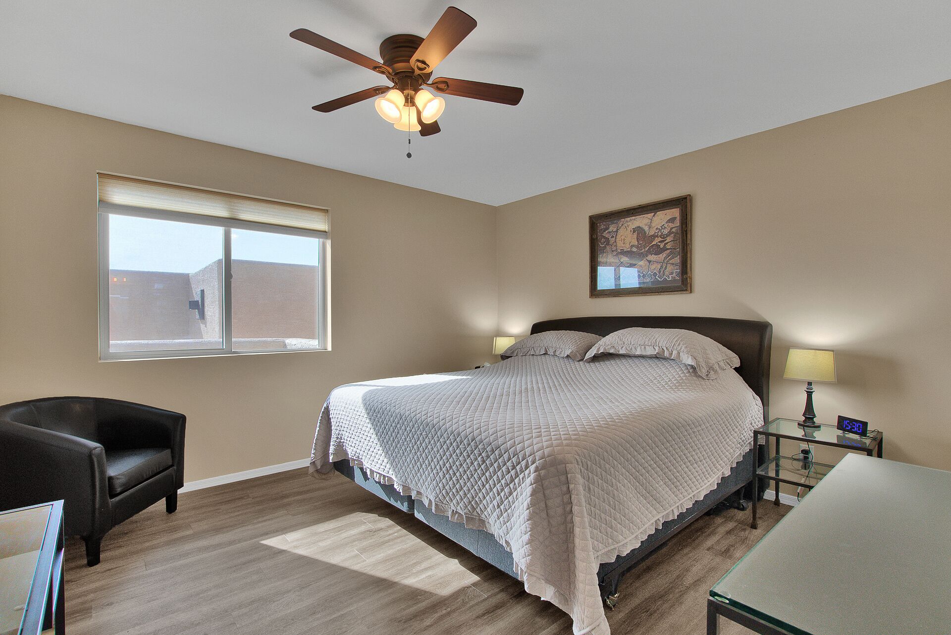 A bedroom with a bed, a chair, a desk, a ceiling fan, a window, and a painting on the wall.