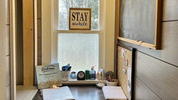 A reception desk with a 'STAY awhile' sign, a welcome sign, a guest book, and a window with a view of trees.