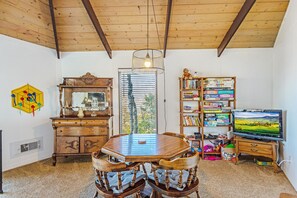 A dining area with a wooden table and chairs, a bookshelf, a television, and a window with blinds.