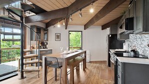 Una cocina moderna con suelos de madera, una zona de comedor con una mesa y sillas de madera, una escalera con un rascador para gatos y vistas al exterior con vegetación.