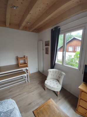 A room with a wooden ceiling, a white armchair, a wooden table, and a window with a view of a house.