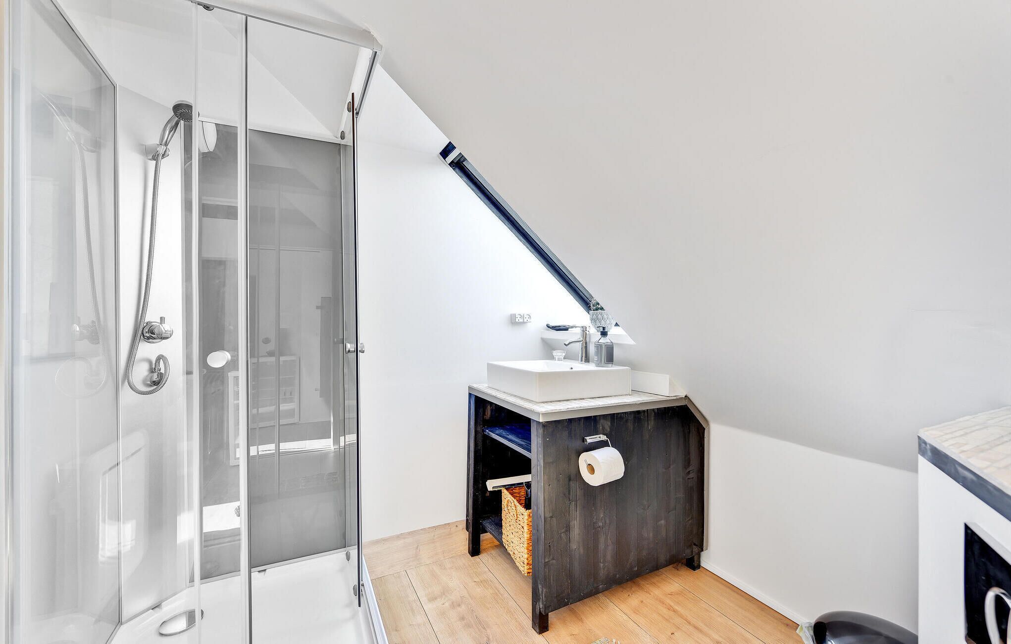 Un baño moderno con ducha de vidrio, un lavamanos blanco con dispensador de toallas y un mueble de madera oscura.