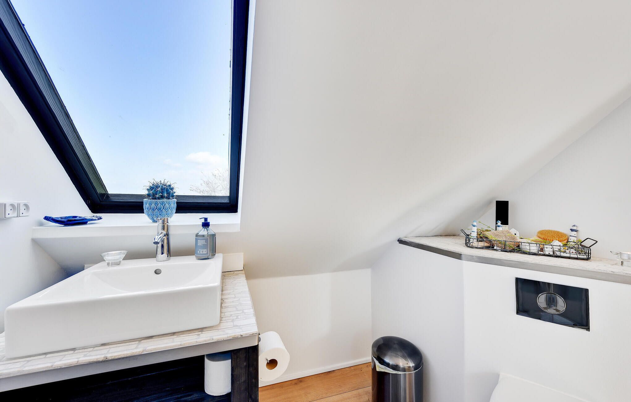 A modern bathroom with a skylight, a white sink, a small shelf with toiletries, and a trash bin.