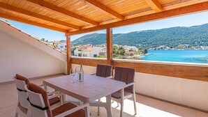 Un balcone con tetto in legno, un tavolo e quattro sedie. Affaccia su una zona costiera con edifici e colline.