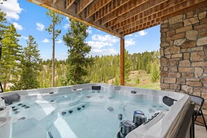A hot tub with a stone wall and wooden ceiling, surrounded by trees and a clear sky.