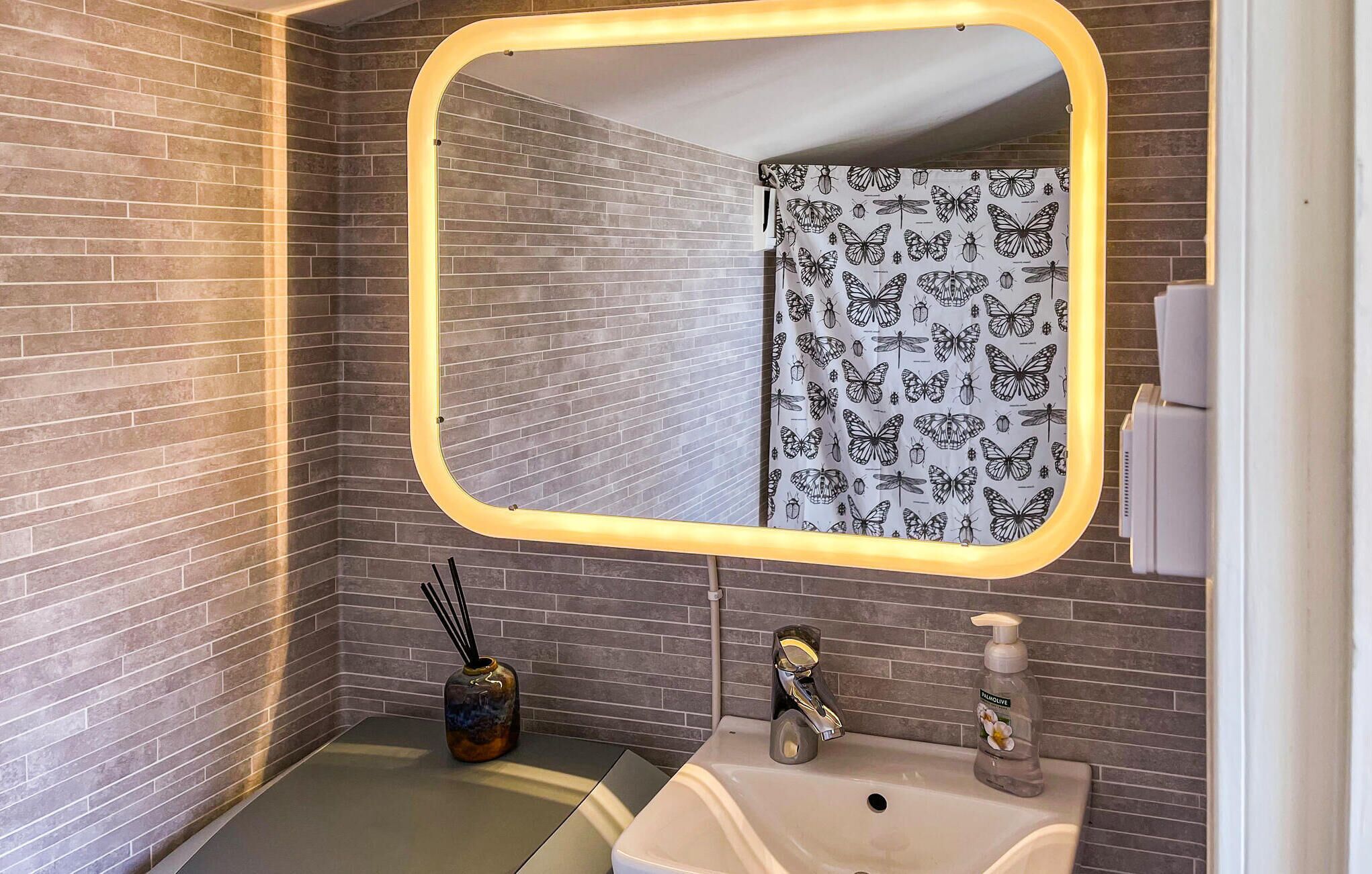Une salle de bain avec un miroir, un lavabo, un rideau de douche orné de motifs de papillons, un vase contenant un diffuseur de parfum à bâtonnets et un distributeur de savon.