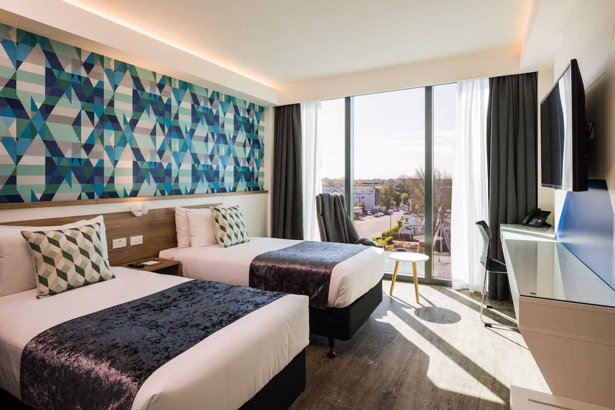 Ein Hotelzimmer mit zwei Betten, einem Schreibtisch, einem Fernseher und einem großen Fenster mit Blick auf die Stadt.