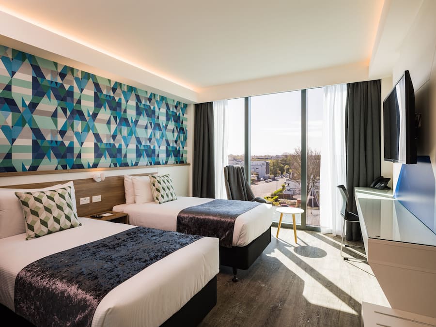Habitación de hotel con dos camas, un escritorio, un televisor y un ventanal con vistas a la ciudad.
