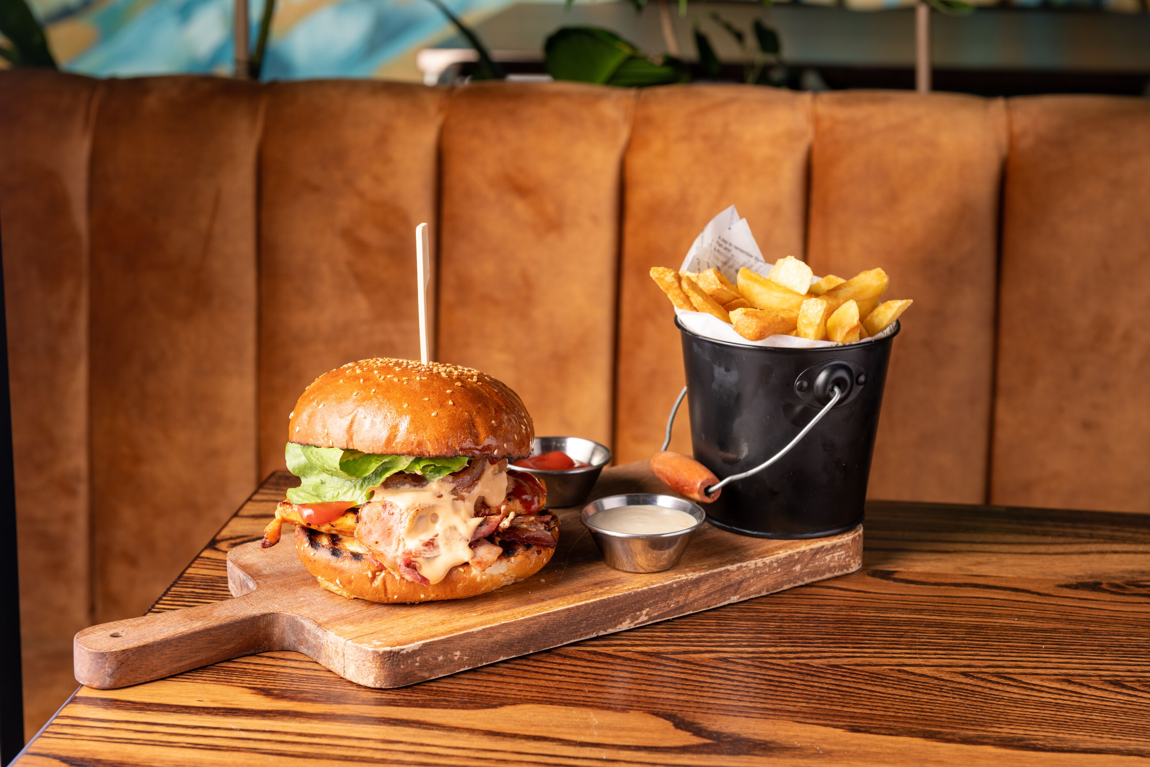 A burger with fries on a wooden board, a drink in a black mug, and a small bowl of sauce on a table.