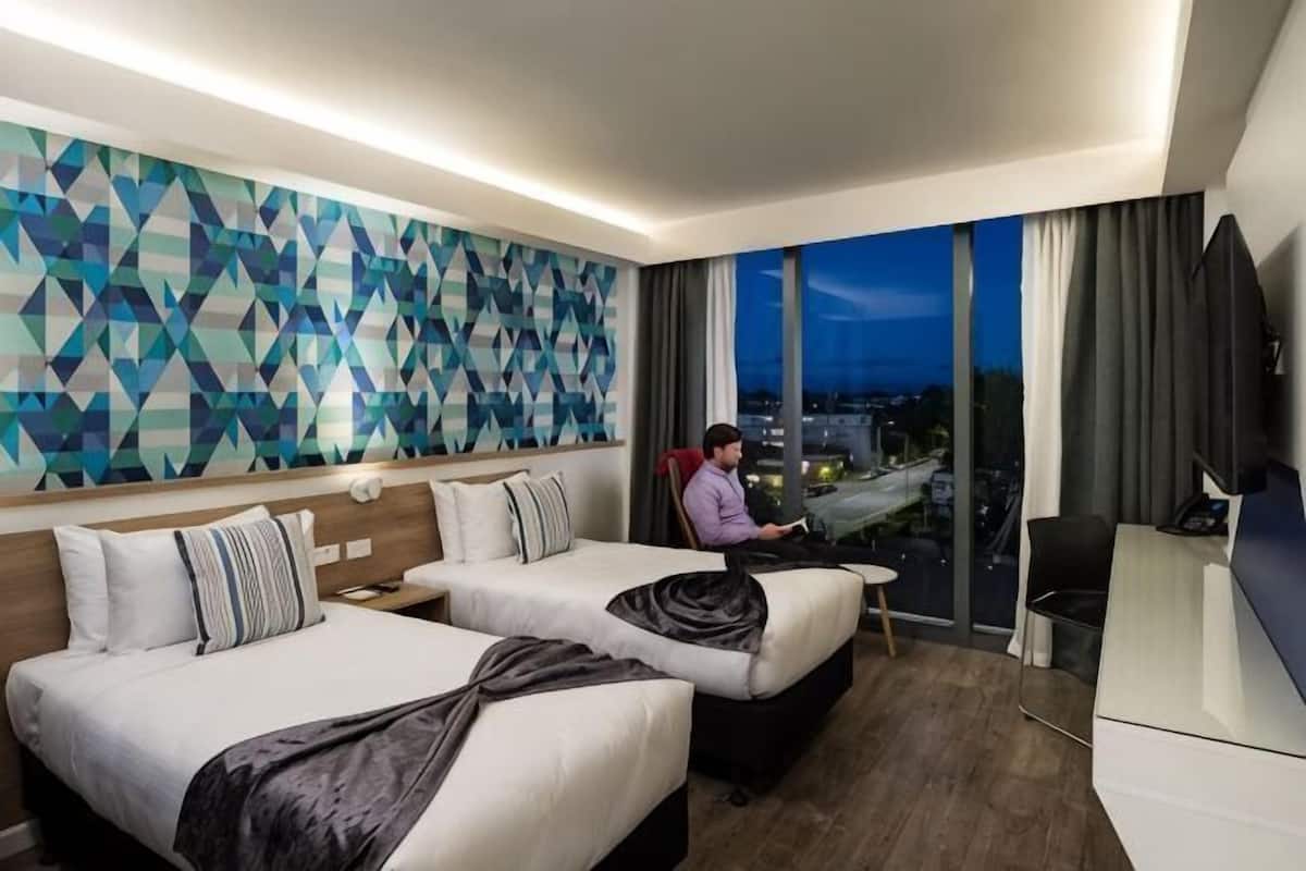 Ein Hotelzimmer mit zwei Betten, einem Schreibtisch, einem Fernseher und einem großen Fenster mit Blick auf die Stadt.