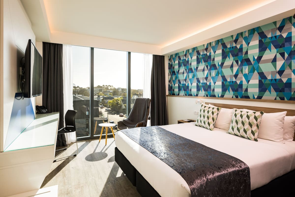 Ein modernes Hotelzimmer mit einem großen Bett, einem Schreibtisch mit Computer, einem Sessel und einem großen Fenster mit Blick auf die Stadt.
