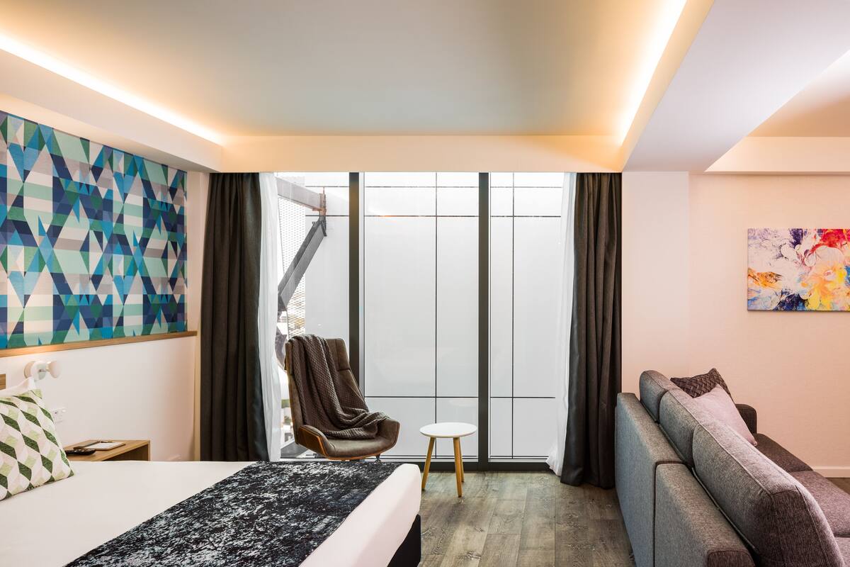 Ein modernes Hotelzimmer mit einem großen Bett, einer Couch, einem Sessel und einem kleinen Tisch. Ein großes Fenster mit Vorhängen, ein geometrisches Wandbild und ein farbenfrohes Gemälde an der Wand.