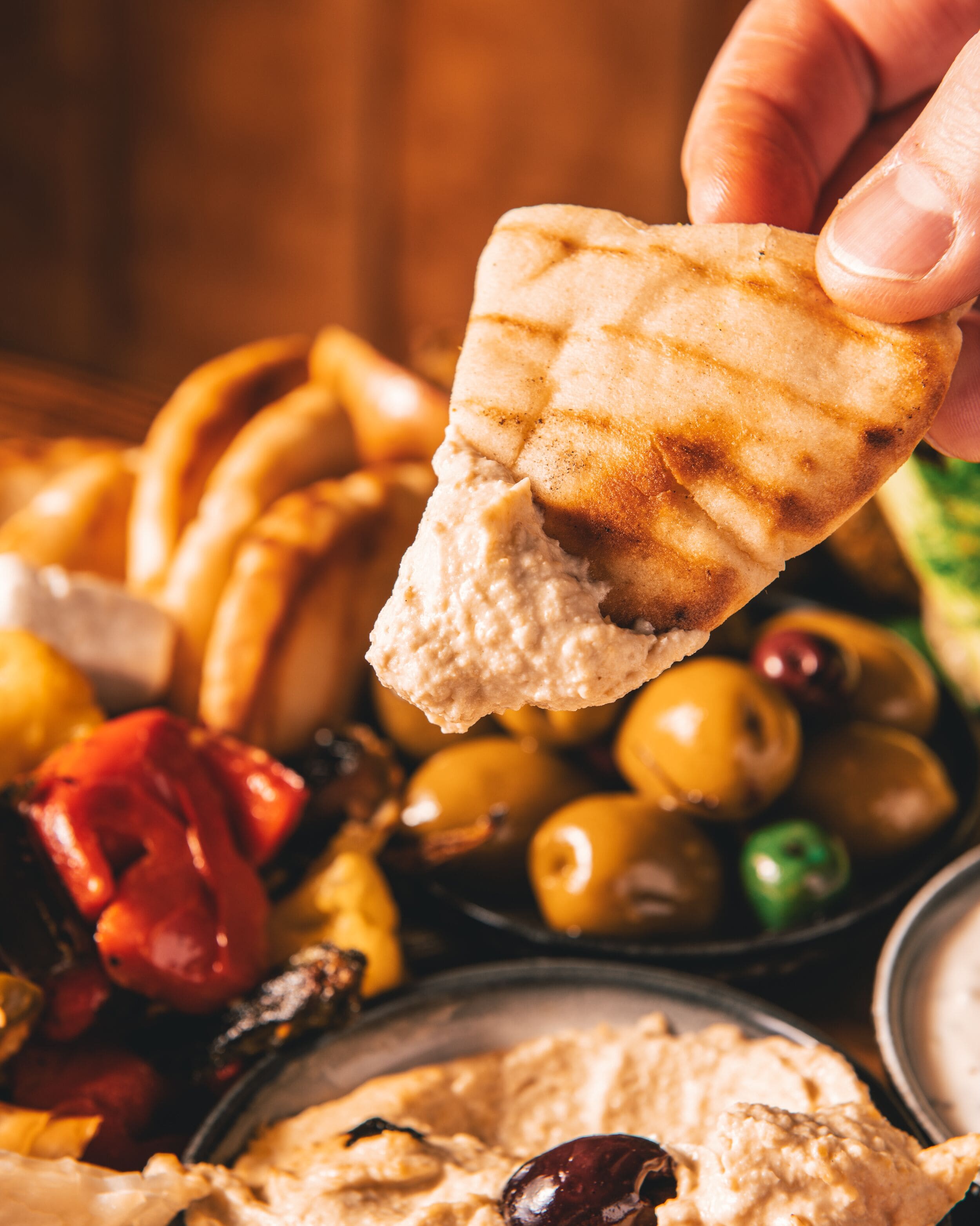 A hand holding a piece of flatbread over a variety of foods including olives, pickles, and a bowl of hummus.