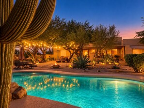 Une piscine à l’eau d’un bleu cristallin, entourée d’un patio aménagé avec des meubles d’extérieur et illuminé de guirlandes lumineuses.