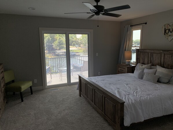 A bedroom with a large bed, a ceiling fan, a nightstand with a lamp, a chair, and a sliding glass door leading to a balcony. - Completely renovated home in Quiet Cove with a Dock! (Sunrise Beach)