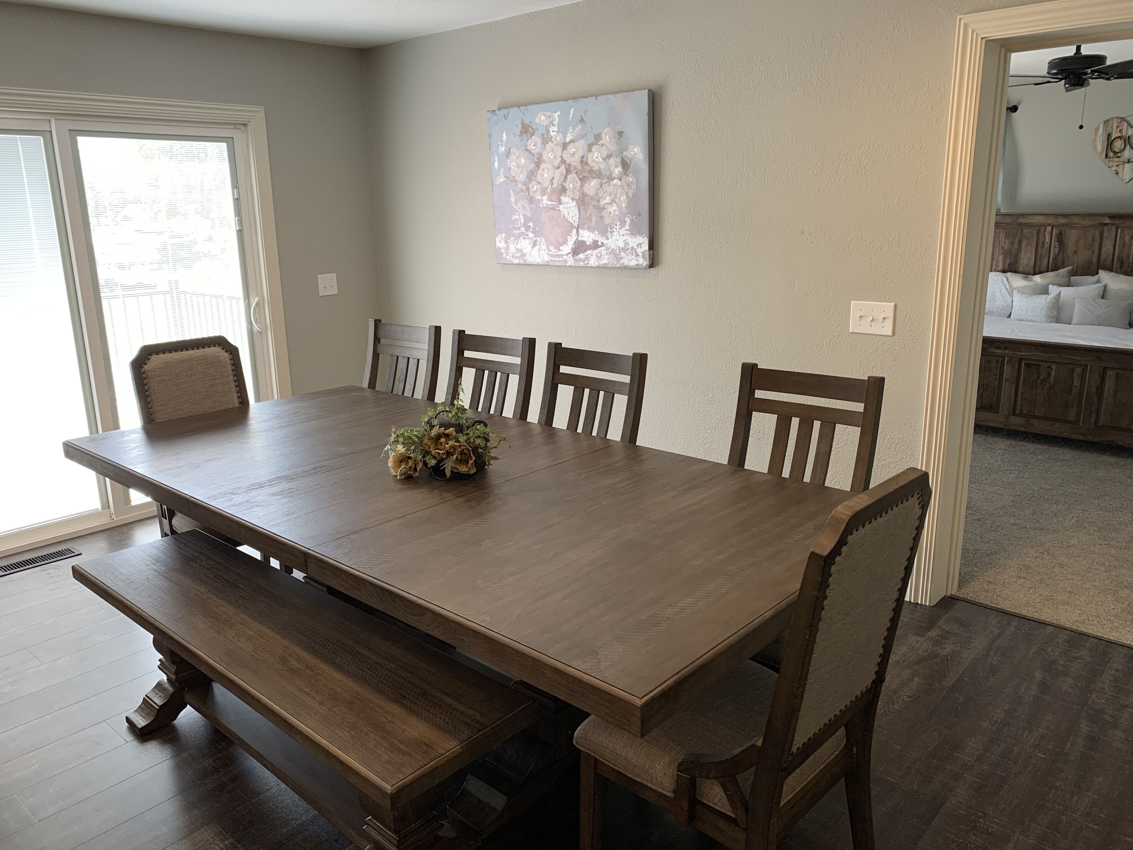 A dining room with a large wooden table, benches, and chairs. There is a painting on the wall and a ceiling fan in the background.