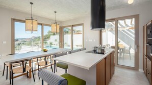 Une cuisine moderne dotée d’un grand îlot central, d’un coin repas et de grandes fenêtres offrant une vue sur une piscine et un espace de détente extérieur.
