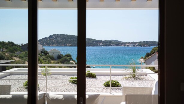 Une vue depuis un balcon, avec une mer bleue et limpide, une colline parsemée de bâtiments et un bâtiment blanc au premier plan.