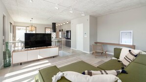 Un salon moderne avec un téléviseur à écran plat, des canapés verts, une table basse en bois et une cuisine en arrière-plan.