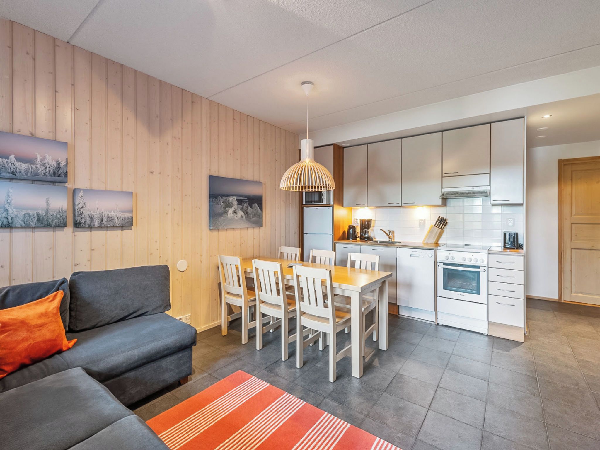Un salon moderne comprenant un canapé, une table à manger et des chaises, une cuisine avec des meubles blancs et des appareils électroménagers, et un lambris en bois sur les murs.