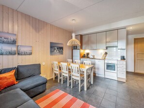 A modern living room with a sofa, dining table, and chairs, a kitchen with white cabinets and appliances, and wooden paneling on the walls.