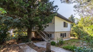 Una casa a due piani con fondamenta in pietra, una scala che conduce al piano superiore e un giardino curato con diverse piante e un palo di legno.