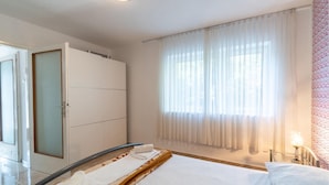 Camera d'albergo con un letto, una grande finestra con tende leggere, un armadio e un bagno visibile attraverso una porta aperta.