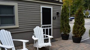 Zwei weiße Adirondack-Stühle vor einem Gebäude mit grauer Fassade und einer schwarzen Tür. Neben den Stühlen stehen je ein Kübel mit immergrünen Bäumen.